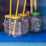 Four mason jar glasses with vibrant lids and straws on a blue shelf.